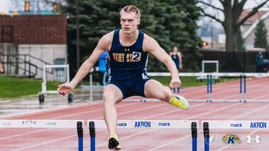 Kent State University track and field athlete Owen Miller, wearing bib number 2, clears a hurdle in rainy conditions during an outdoor track meet at Akron. He wears the navy blue and gold Kent State uniform. Hurdles branded with the Akron and UCS logos line the track behind him. The Kent State Athletics logo and Under Armour wordmark appear in the lower right corner.
