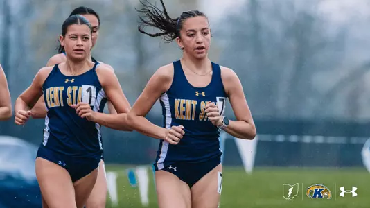 Kent State University distance runners Nora Salem (right) and Emerson Greene (left) compete side by side during an outdoor track meet in rainy conditions, wearing navy blue and gold Kent State uniforms with bib numbers 7 and 1. A third runner is visible behind them. The Kent State Athletics logo and Under Armour wordmark appear in the lower right corner.