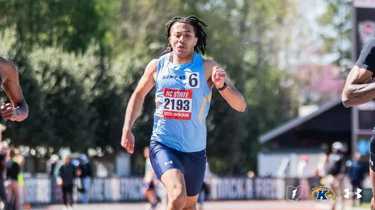 Kent State University track and field athlete Chaz Sakala, wearing bib number 2193 and a light blue Kent State uniform, races at the NC State Raleigh Relays outdoor track meet. The athlete wears the meet's adidas race bib over his uniform. Competitors and spectators are visible on either side. The Kent State Athletics logo and Under Armour wordmark appear in the lower right corner.