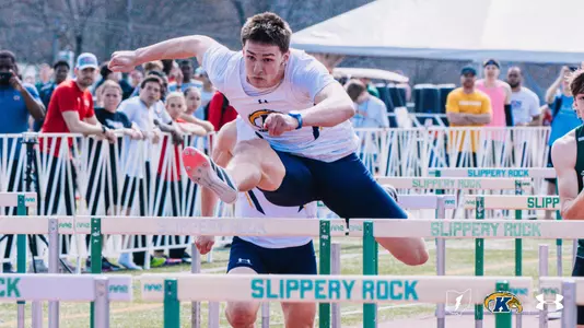 Kent State University track and field athlete Linkon Tyrrell, wearing a white Kent State uniform with the Golden Flashes logo, powers over a hurdle at the Slippery Rock outdoor track meet, with a large crowd of spectators watching from behind a barrier. Hurdles branded with the Slippery Rock name line the track. The Kent State Athletics logo and Under Armour wordmark appear in the lower right corner.