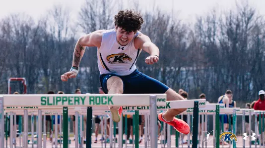 Kent State University track and field athlete Mason Murphy, wearing a white Kent State uniform with the Golden Flashes logo, clears a hurdle at the Slippery Rock outdoor track meet, with rows of hurdles and other competitors visible in the background. The Kent State Athletics logo and Under Armour wordmark appear in the lower right corner.