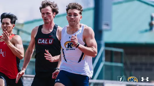Kent State University track and field athlete Ethan Pater, wearing a white Kent State uniform featuring the Golden Flashes logo, races alongside a competitor in a black California University of Pennsylvania uniform during an outdoor meet at Slippery Rock. A third runner in a red uniform is visible to the left. The Kent State Athletics logo and Under Armour wordmark appear in the lower right corner.