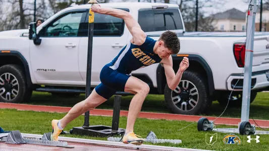 Kent State University track and field athlete Jack Dillen drives off the starting block during an outdoor track meet at Akron, wearing a navy blue and gold Kent State uniform. Starting blocks and pole vault equipment are visible on the track, with a white pickup truck in the background. The Kent State Athletics logo and Under Armour wordmark appear in the lower right corner.