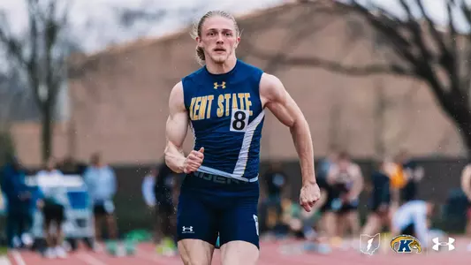 Kent State University track and field athlete Logan Doyle, wearing bib number 8, races in rainy conditions during an outdoor track meet at Akron. He wears the navy blue and gold Kent State uniform with an Under Armour logo. Spectators are blurred in the background. The Kent State Athletics logo and Under Armour wordmark appear in the lower right corner.