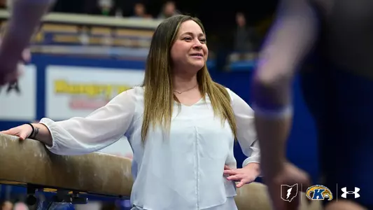 A Kent State gymnastics coach in a white blouse leans against the balance beam on the competition floor, smiling warmly as she watches her athletes during a meet. Gymnasts are visible in the blurred foreground and background, with the arena's blue and gold signage visible behind her.