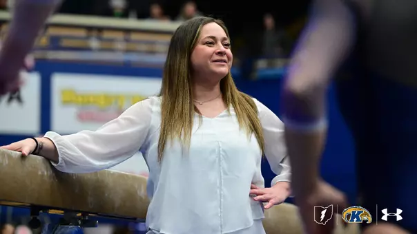 A Kent State gymnastics coach in a white blouse leans against the balance beam on the competition floor, smiling warmly as she watches her athletes during a meet. Gymnasts are visible in the blurred foreground and background, with the arena's blue and gold signage visible behind her.