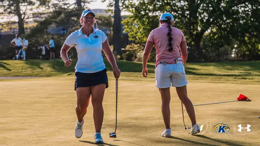 Kent State golfer Aryn Matthews grins and pumps her fist as she walks off the green during the Therese Hession Buckeye Invitational at Ohio State University Golf Club. Matthews is wearing a white Kent State polo, navy skort and blue cap, carrying her putter at her side. An opposing player stands to her right, facing away. Warm late-afternoon sunlight illuminates the green.