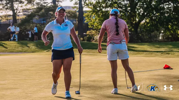Kent State golfer Aryn Matthews grins and pumps her fist as she walks off the green during the Therese Hession Buckeye Invitational at Ohio State University Golf Club. Matthews is wearing a white Kent State polo, navy skort and blue cap, carrying her putter at her side. An opposing player stands to her right, facing away. Warm late-afternoon sunlight illuminates the green.