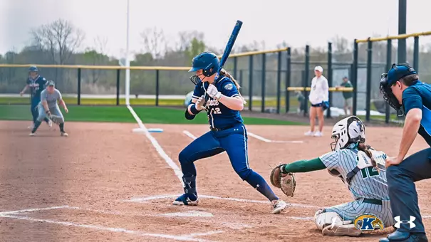 Kent State softball player Ashlyn Porter, wearing jersey number 12, stands in the batter's box in a full swing stance during a game. She is dressed in a navy Kent State uniform with a blue batting helmet and face guard, and white batting gloves. The opposing team's catcher in a white and green pinstripe uniform crouches to her right, with the home plate umpire positioned behind. In the background, an opposing baserunner can be seen rounding a base on the left side of the field. The Kent State Golden Flashes and Under Armour logos appear in the bottom right corner.