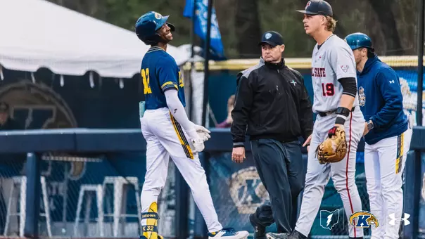 Kent State Golden Flashes baseball player wearing No. 32 in a navy blue jersey and white pants smiles broadly while standing near first base, with an umpire and an opposing player in a white jersey with No. 19 visible in the background, and a Kent State base coach partially visible at right.