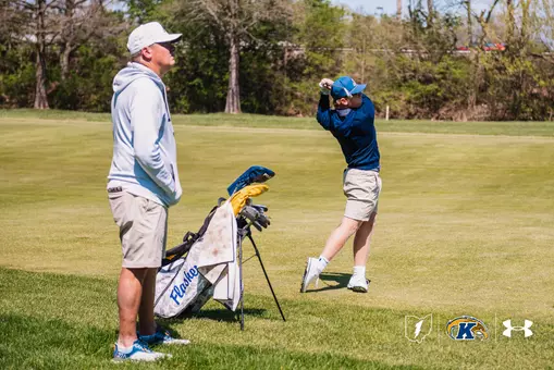 Kent State men's golfer Liam Curtis completes his follow-through on an iron shot in the fairway as head coach Jon Mills watches from behind the team bag. Mills wears a white Kent State cap and hoodie, khaki shorts and blue shoes. Curtis wears a blue Kent State cap, navy long-sleeve pullover, khaki shorts and white golf shoes, with his club finishing high over his left shoulder. The camo-pattern Kent State Flashes stand bag sits between them on the fairway under bright spring sunlight.