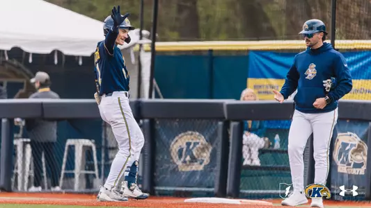 A Kent State Golden Flashes baseball player in a navy jersey and white pants smiles and waves to the dugout while standing on first base, with a Kent State base coach in a navy Golden Flashes pullover and white pants looking on beside him, and the Kent State 'K' logo visible on the outfield wall behind them.
