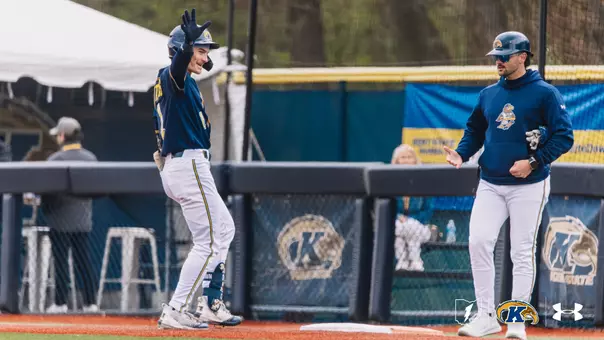 A Kent State Golden Flashes baseball player in a navy jersey and white pants smiles and waves to the dugout while standing on first base, with a Kent State base coach in a navy Golden Flashes pullover and white pants looking on beside him, and the Kent State 'K' logo visible on the outfield wall behind them.