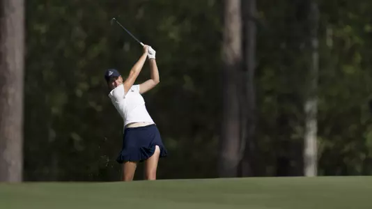 Kent State junior Veronika Kedronová hits an approach shot from the fairway during the second round of the Augusta National Women's Amateur at Champions Retreat Golf Club, wearing a white polo and navy skort with pine trees lining the background.