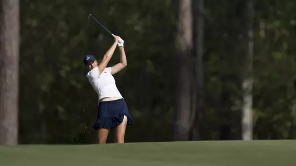 Kent State junior Veronika Kedronová hits an approach shot from the fairway during the second round of the Augusta National Women's Amateur at Champions Retreat Golf Club, wearing a white polo and navy skort with pine trees lining the background.