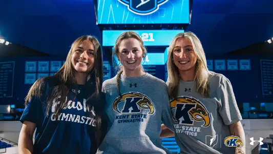 Charlie Behner, Heidi Schultz, and Jersey Bingman are smiling and wearing Kent State Gymnastics shirts. Behind them is a well-lit sports venue with banners and logos visible. The setting appears to be inside a gymnasium or sports arena.