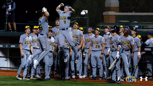 A group of baseball players in gray Kent State uniforms stand in a dugout. Two players are jumping and giving each other a high-five. The dugout is lively, with players holding bats and looking on. The Kent State logo is visible on the uniforms and equipment.