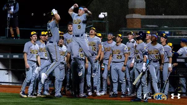 A group of baseball players in gray Kent State uniforms stand in a dugout. Two players are jumping and giving each other a high-five. The dugout is lively, with players holding bats and looking on. The Kent State logo is visible on the uniforms and equipment.