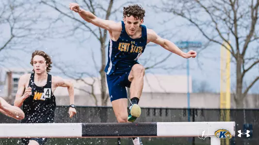 Kent State University track and field athlete Owen Kelly clears a steeplechase hurdle in rainy conditions during an outdoor track meet at Akron, wearing a navy blue and gold Kent State uniform. A competitor in a black uniform trails behind him. The Kent State Athletics logo and Under Armour wordmark appear in the lower right corner.