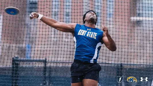 Kent State University track and field athlete Desmond Boyd releases a discus during an outdoor meet at Akron in rainy conditions, wearing a navy blue and gold Kent State uniform. A safety cage surrounds the throwing circle, with a brick building visible in the background. The Kent State Athletics logo and Under Armour wordmark appear in the lower right corner.