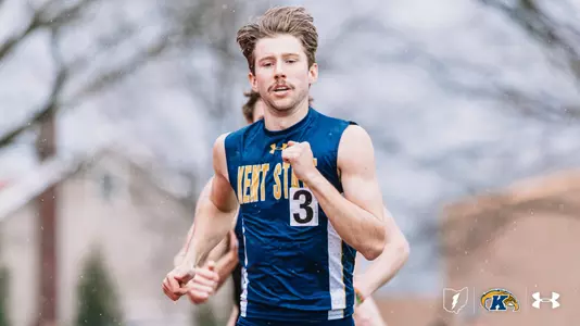 Kent State University track and field athlete Jack Curtis, wearing bib number 3, competes in rainy conditions during an outdoor track meet at Akron, dressed in a navy blue and gold Kent State uniform. A second Golden Flashes runner is visible behind him. The Kent State Athletics logo and Under Armour wordmark appear in the lower right corner.