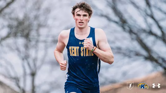 Kent State University track and field athlete Caleb Wilcox competes in rainy conditions during an outdoor track meet at Akron, wearing a navy blue and gold Kent State uniform. Rain is visible throughout the frame against a backdrop of bare trees. The Kent State Athletics logo and Under Armour wordmark appear in the lower right corner.