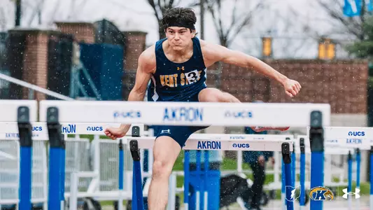 Kent State University track and field athlete Beau Harkelroad, wearing bib number 3 and a navy blue and gold Kent State uniform, clears a hurdle in rainy conditions during an outdoor track meet at Akron. Hurdles branded with the Akron and UCS logos line the track. The Kent State Athletics logo and Under Armour wordmark appear in the lower right corner.