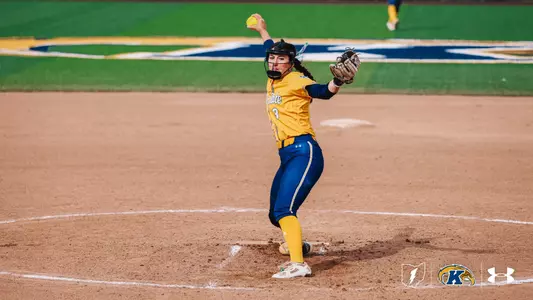 "Kent State softball pitcher Brynn Libler, wearing jersey number 3, winds up to deliver a pitch during a home game. She is dressed in a gold and navy Kent State uniform with gold pants, a black helmet with face guard, and a brown fielding glove. A yellow softball is visible above her head at the top of her wind-up. She is positioned on the pitcher's mound with the Kent State 'K' logo visible on the outfield grass in the background. The Ohio Flash, Kent State Golden Flashes, and Under Armour logos appear in the bottom right corner."