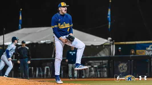 A Kent State Golden Flashes pitcher in a navy blue 'Kent State' Under Armour jersey, white pants, and gold-brimmed cap pumps his fist and shouts in celebration on the mound under the lights, with an opposing baserunner and the Kent State dugout visible in the background.