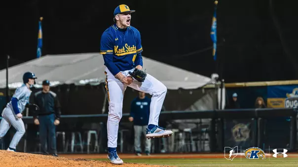 A Kent State Golden Flashes pitcher in a navy blue 'Kent State' Under Armour jersey, white pants, and gold-brimmed cap pumps his fist and shouts in celebration on the mound under the lights, with an opposing baserunner and the Kent State dugout visible in the background.