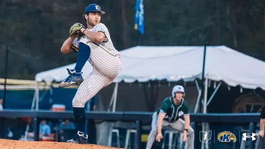 A Kent State Golden Flashes pitcher in a white pinstripe Under Armour jersey, navy socks, and a navy Golden Flashes cap winds up in a high leg kick on the mound, with an opposing baserunner in a green jersey taking a lead in the background.