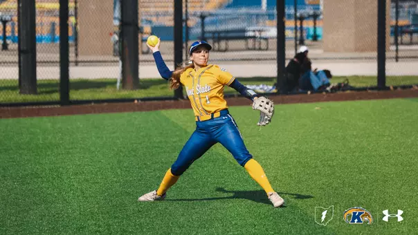 "Kent State softball outfielder Brooke Aberle, wearing jersey number 1, winds up to make a throw from the outfield during a home game. She is dressed in a gold Kent State jersey with navy pants, gold knee-high socks, a navy long-sleeve undershirt, a blue cap, and a tan fielding glove. A yellow softball is visible in her raised throwing hand. She is positioned in a wide stance on the green turf outfield. The stadium seating and fencing are visible in the blurred background. The Ohio Flash, Kent State Golden Flashes, and Under Armour logos appear in the bottom right corner."