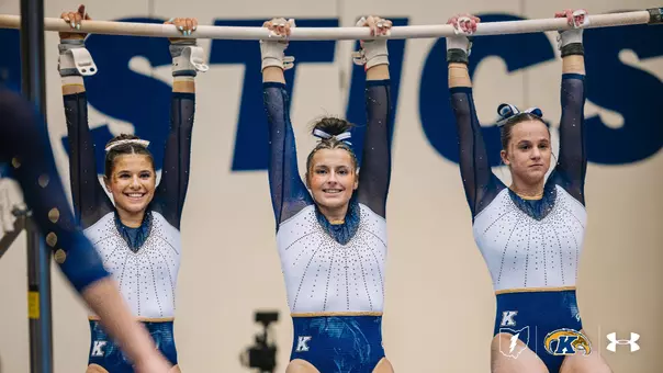 Three Kent State Golden Flashes gymnasts in navy, white, and gold rhinestone-embellished leotards hang side by side from the uneven bars, smiling, with the word 'Gymnastics' visible on the wall behind them and the Kent State lightning bolt, Golden Flashes, and Under Armour logos in the corner.