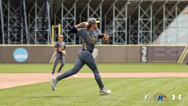 A Kent State Golden Flashes infielder in a gray 'Kent State Golden Flashes' Under Armour jersey and gray pants fires a throw across the diamond, with a teammate visible in the background and the NCAA logo on the outfield wall, during a game at a stadium with exposed steel bleacher framework.