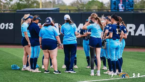 "The Kent State softball team huddles together on the outfield turf during a game. Players and coaches are dressed in a mix of light blue and navy Kent State uniforms. Coaches address the group during what appears to be a timeout or pregame meeting. A light blue fielding glove rests on the ground to the left. The outfield wall with championship year markings is visible in the background, along with a video scoreboard. The Ohio Flash, Kent State Golden Flashes, and Under Armour logos appear in the bottom right corner."