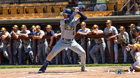 Kent State Golden Flashes batter wearing No. 8 stands in his stance at home plate in a gray 'Kent State Golden Flashes' Under Armour jersey and gray pants, with the entire Kent State dugout leaning on the railing watching in the background and an opposing catcher crouched at right.