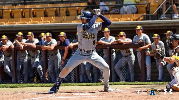 Kent State Golden Flashes batter wearing No. 8 stands in his stance at home plate in a gray 'Kent State Golden Flashes' Under Armour jersey and gray pants, with the entire Kent State dugout leaning on the railing watching in the background and an opposing catcher crouched at right.