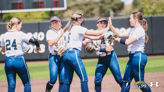 Five Kent State softball players celebrate together on the field during a game, exchanging high-fives in a group. The players are wearing white pinstripe Kent State uniforms with blue pants. Number 24 is visible on the player at far left, and the center player is wearing a catcher's helmet pushed up on her head and holding a fielding glove. A scoreboard showing 8-5 is visible in the background, along with the outfield wall displaying championship years. The Ohio Flash, Kent State Golden Flashes, and Under Armour logos appear in the bottom right corner."