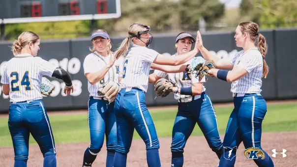 Five Kent State softball players celebrate together on the field during a game, exchanging high-fives in a group. The players are wearing white pinstripe Kent State uniforms with blue pants. Number 24 is visible on the player at far left, and the center player is wearing a catcher's helmet pushed up on her head and holding a fielding glove. A scoreboard showing 8-5 is visible in the background, along with the outfield wall displaying championship years. The Ohio Flash, Kent State Golden Flashes, and Under Armour logos appear in the bottom right corner."