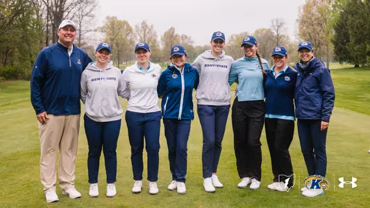 The Kent State Dr. Paula Treckel women's golf team poses for a group photo on the course at Firestone Country Club ahead of the 2026 MAC Championship. Eight people stand in a row with arms around each other, smiling at the camera. From left: head coach Casey VanDamme in a navy quarter-zip and Kent State cap, Aryn Matthews in a gray Kent State hoodie, Gracie Larsen in a white pullover, Leon Takagi in a navy zip jacket, Veronika Kedronová in a gray Kent State hoodie, Petra Babicová in a light blue quarter-zip, Isabella Goyette in a navy polo, and assistant coach Manuela Carbajo Ré in a navy rain jacket. Trees and fairway extend behind them under overcast skies.