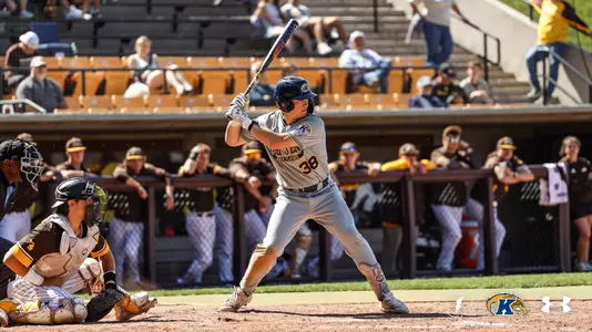Kent State Golden Flashes batter wearing No. 38 stands ready in his stance at home plate in a gray 'Kent State Golden Flashes' Under Armour jersey and gray pants, with an opposing catcher and umpire at left, the opposing dugout lined with players watching in the background, and fans in the bleachers behind them.