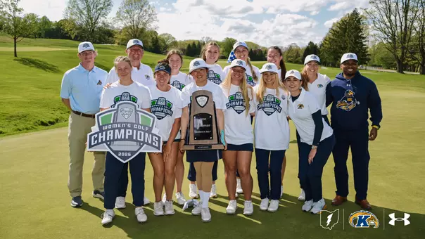 The Kent State Dr. Paula Treckel women's golf team poses on a manicured green at Firestone Country Club's Fazio Course after winning the 2026 Mid-American Conference Women's Golf Championships. Twelve people stand together in two loose rows, smiling at the camera. The player at front center holds the wooden MAC championship trophy, while another player at front left holds a large 2026 MAC Women's Golf Champions shield-shaped sign. Most of the team wears matching white commemorative champions t-shirts and white champions hats, with several in navy Kent State polos. Behind them, a sweeping fairway slopes down to mature trees under a blue sky scattered with white clouds. The Ohio-shaped Golden Flashes logo, Kent State athletic mark, and Under Armour logo appear in the bottom right corner.