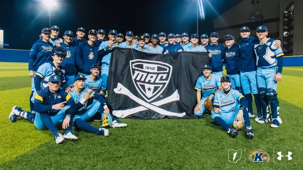 The Kent State baseball team gathers on the field under the lights for a group photo, holding a large black Mid-American Conference flag with crossed bats, wearing light blue 'Flashes' Under Armour uniforms and navy Kent State Baseball jackets, celebrating a victory.