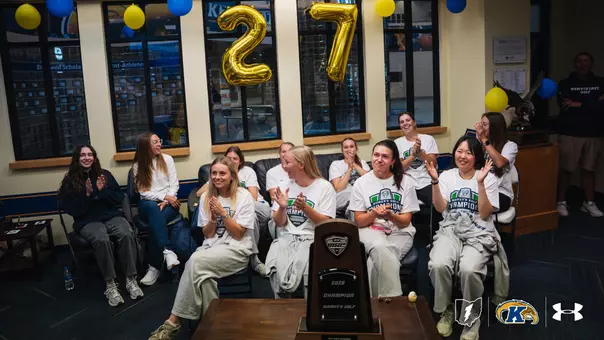 Kent State women's golf team members clap and celebrate in a lounge decorated with blue and gold balloons and large gold '27' balloons, gathered around the 2026 MAC Women's Golf Championship trophy displayed in the foreground, with several players wearing white MAC Women's Golf Champions t-shirts and the Kent State lightning bolt, Golden Flashes, and Under Armour logos in the corner.