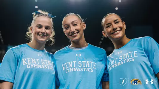 Heidi Schultz, Jersey Bingman, and Charlie Behner wearing blue "Kent State Gymnastics" shirts are smiling. The Under Armour logo and Kent State University logos are visible at the bottom. The background is dark and out of focus.
