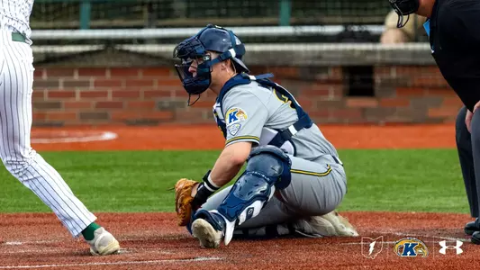 A baseball player wearing a gray and blue uniform with protective gear is crouched behind home plate. The uniform features a "K" emblem, indicating Kent State University. Another player in a striped uniform is standing at the plate. The field is a mix of green turf and red dirt. Logos for the Mid-American Conference and Under Armour are visible in the bottom right corner.