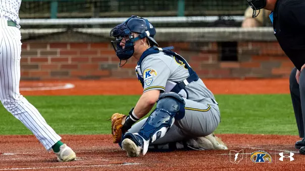 A baseball player wearing a gray and blue uniform with protective gear is crouched behind home plate. The uniform features a "K" emblem, indicating Kent State University. Another player in a striped uniform is standing at the plate. The field is a mix of green turf and red dirt. Logos for the Mid-American Conference and Under Armour are visible in the bottom right corner.