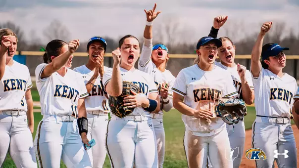 A group of Kent State University softball players in white and navy Under Armour uniforms celebrate enthusiastically on the field. Several players are shouting and raising their fists in the air, with some holding softball gloves. One player's uniform is visibly dirty from sliding. The photo is taken outdoors on an overcast spring day with bare trees visible in the background.