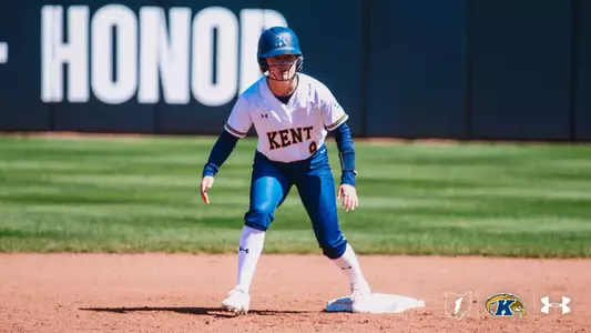 Kent State University softball player Kelsey Salopek (#9) crouches in an athletic ready stance on second base, preparing to run. She is wearing a white Kent State Under Armour jersey, blue pants, white socks, and a blue batting helmet with a face guard. The outfield grass and a dark outfield wall with the word 'HONOR' are visible in the background. The Ohio outline, Kent State Golden Flashes, and Under Armour logos appear in the bottom right corner.