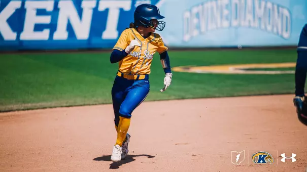 Kent State University softball player Brooke Aberle (#1) runs the bases in full stride, wearing the team's gold alternate Under Armour jersey, blue pants, gold socks, and a navy batting helmet with a face guard. She is wearing batting gloves and pumping her fists as she runs. The Kent State 'KENT' signage and a 'Devine Diamond' banner are visible on the outfield wall in the background. The Ohio outline, Kent State Golden Flashes, and Under Armour logos appear in the bottom right corner.